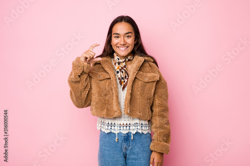 Young mixed race indian woman wearing a short sheepskin coatholding something little with forefingers, smiling and confident.