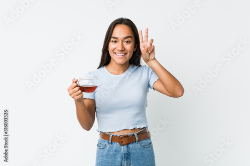 Young mixed race indian holding a tea cup showing victory sign and smiling broadly.