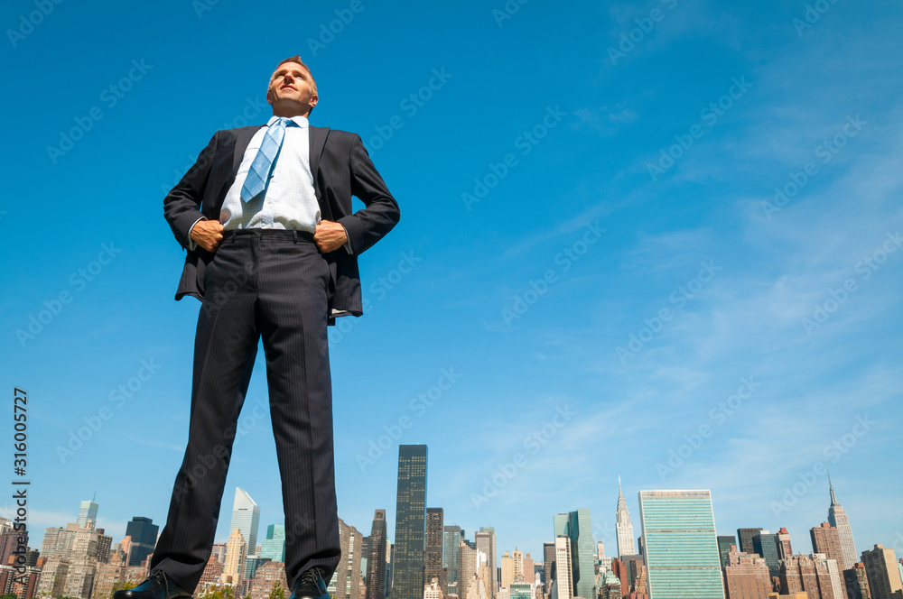Giant businessman standing tall in dark suit above the city skyline - obrazy, fototapety, plakaty 