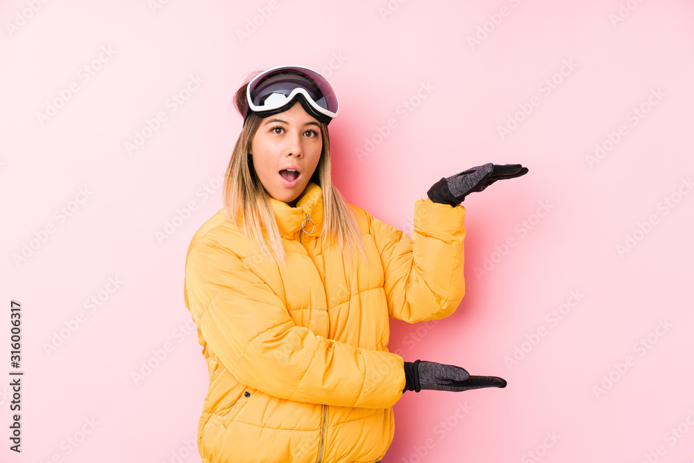 Young caucasian woman wearing a ski clothes in a pink background shocked and amazed holding a copy space between hands. Young caucasian woman wearing a ski clothes in a pink background shocked and amazed holding a copy space between hands.