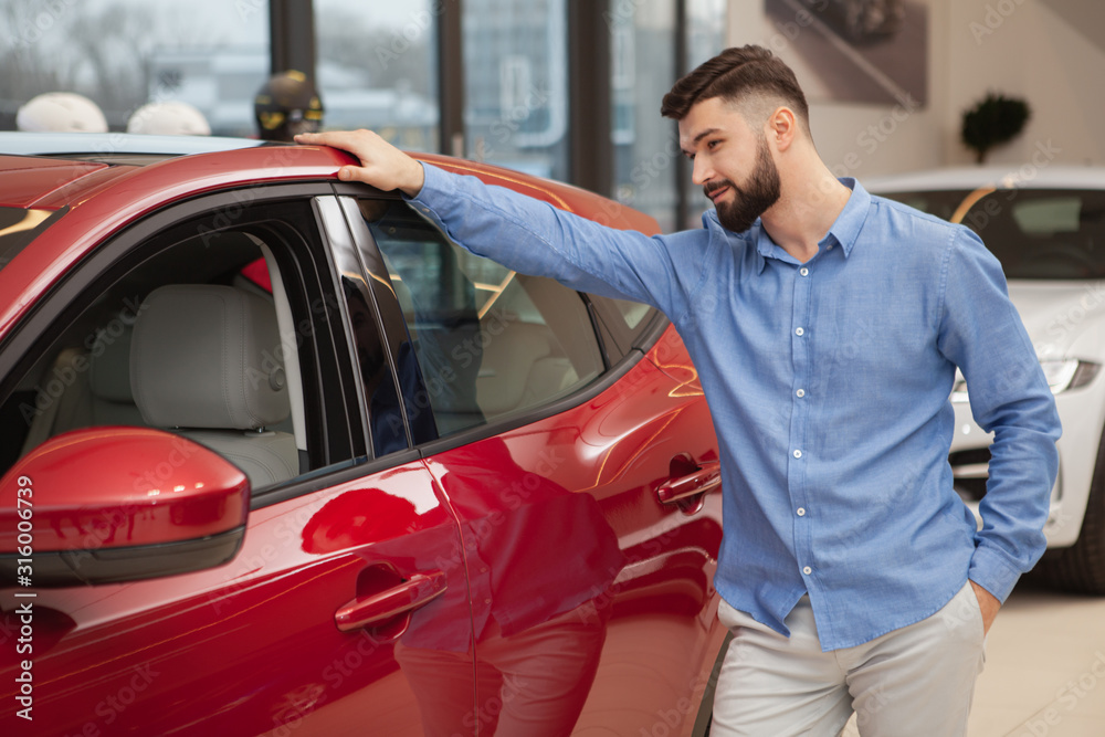 Handsome bearded man examining red automobile at car dealership. Male customer buying new auto