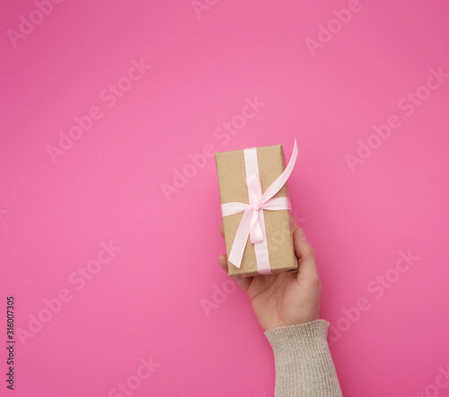 Fototapeta Naklejka Na Ścianę i Meble -  female hand holds a brown box with a pink bow on a pink background