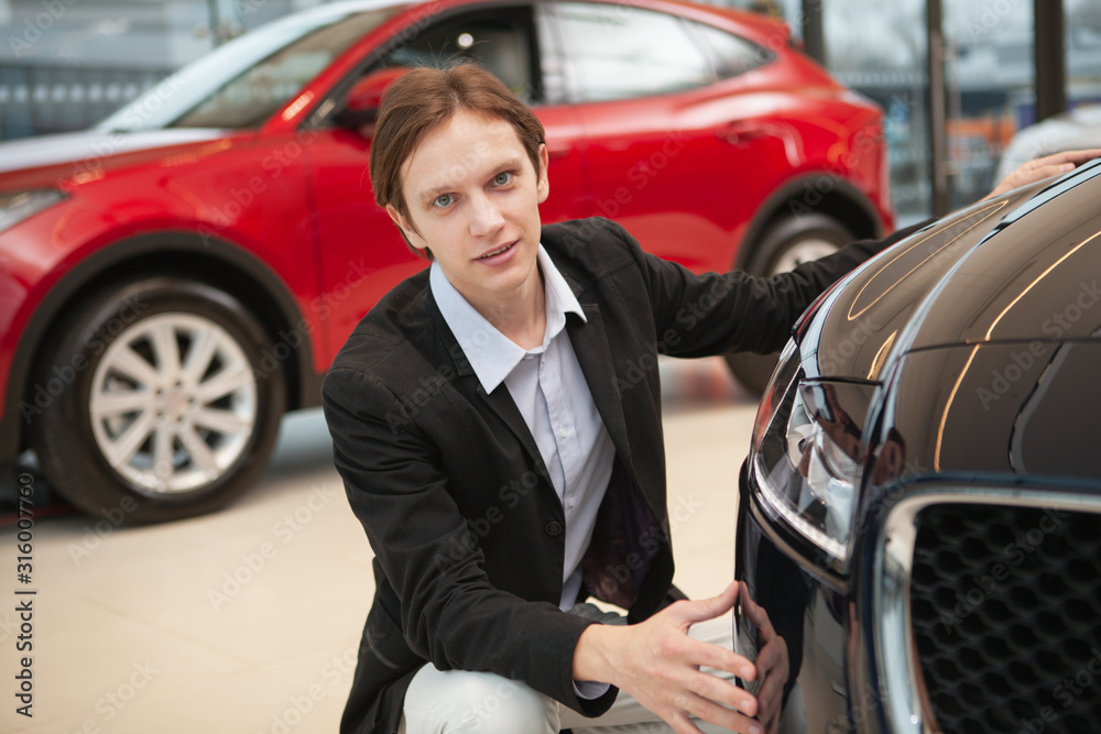 Young elegant man looking to the camera confidently while examining cars on sale at the dealership