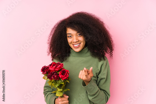 Young afro woman holding a roses isolated Young afro woman holding a rosespointing with finger at you as if inviting come closer.