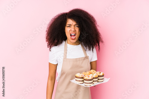 Young afro pastry maker woman holding a cupcakes isolatedYoung afro baker woman screaming very angry and aggressive.