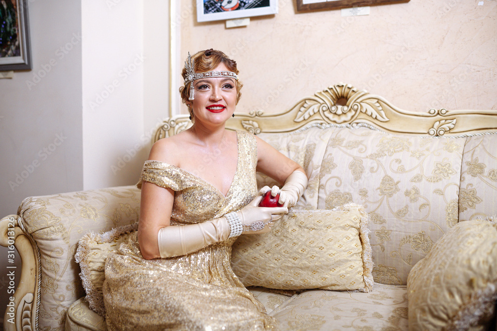 Gatsby style 30s woman in evening dress with a diadem on her head and long gloves. sitting on the couch, holding an apple in his hands and smiling