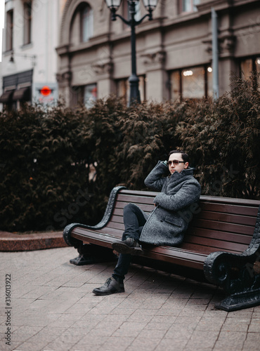 Lone male traveler in a gray coat and glasses sits on a bench in the city.