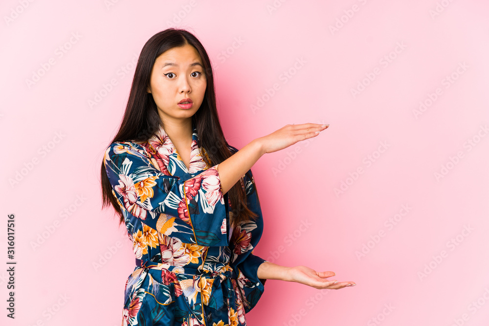 Young chinese woman wearing a kimono pajama isolated shocked and amazed holding a copy space between hands. Young chinese woman wearing a kimono pajama isolated shocked and amazed holding a copy space between hands.