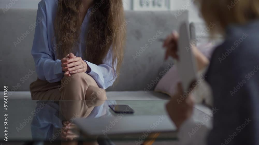 Rack focus of unrecognizable female therapist gesturing while explaining problem solution to young female patient sitting on sofa
