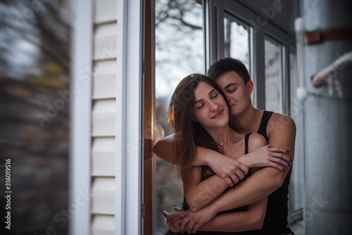 A loving young couple hugs in the window of their balcony terrace in the apartment. Young man laughs with a beautiful woman at home. Lovers kiss, passionately spend time with each other. Lifestyle © farmuty
