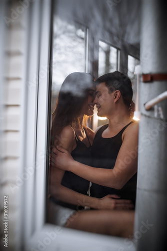 A loving young couple hugs in the window of their balcony terrace in the apartment. Young man laughs with a beautiful woman at home. Lovers kiss  passionately spend time with each other. Lifestyle