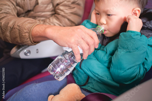 Asian 2 - 3 years old toddler boy child holding his aching ear and drinking water from bottle during flight on airplane. Little kid feeling earache on airplane, Flying with children, Selective focus photo