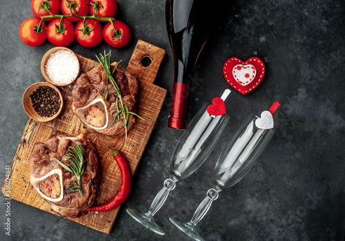 two grilled steaks, with tomatoes and spices with a bottle of wine and glasses, various red hearts on a stone background. dinner for two on valentine's day