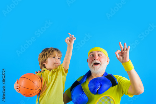Family time. Grandfather and grandson shows sign ok. Family sport. Old man with boxing gloves. Boy holds basketball ball. Grandfather and son workout. Boxing gloves. Sporting. Basketball. Sport game.