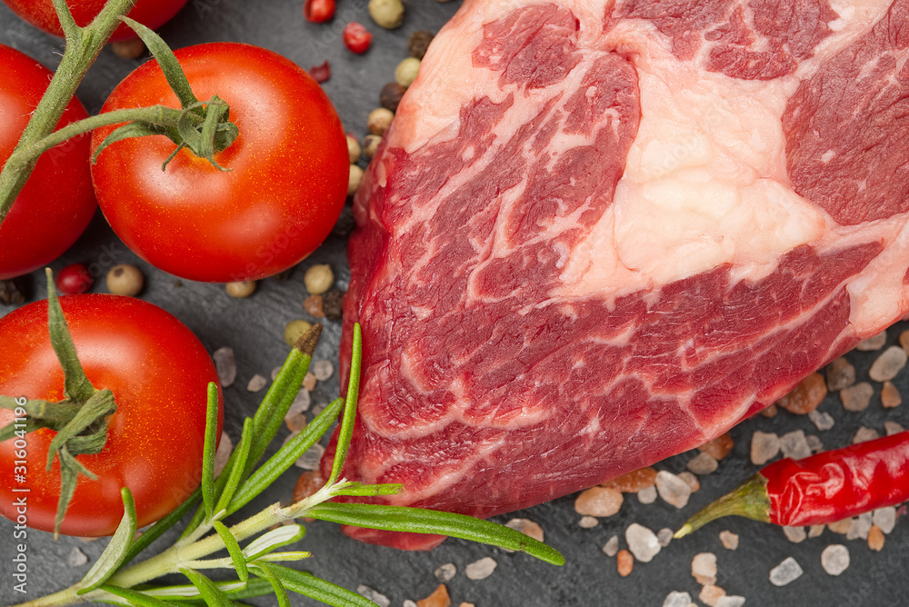 Raw rib eye steak with spices and vegetables. Ingredients for restaurant meal. Fresh meat, salt, rosemary, thyme, chilli, cherry tomatoes, garlic on black stone. Food background. Raw rib eye steak with spices and vegetables. Ingredients for restaurant meal. Fresh meat, salt, rosemary, thyme, chilli, cherry tomatoes, garlic on black stone. Food background.