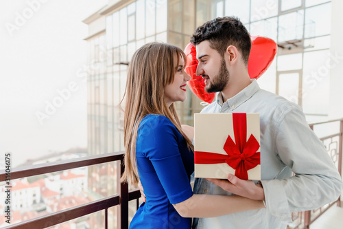 man and woman with gift and red heart shaped balloons on balcony at home
