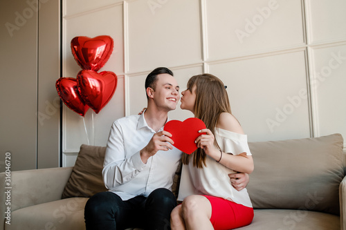 man and woman with red heart shaped valentine card and balloons at home on couch