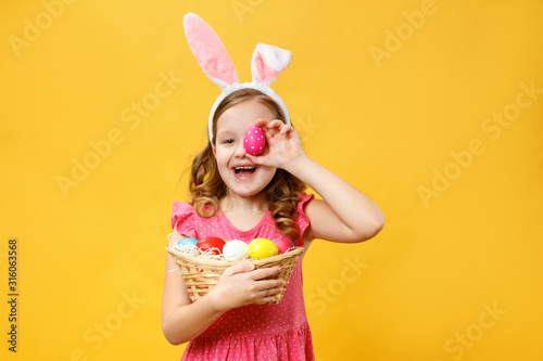 Happy child in the ears of a bunny holds an Easter egg and a basket. Portrait of a little girl on a yellow background