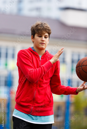 Cute boy in red t shirt plays basketball on city playground. Active teen enjoying outdoor game with orange ball. Hobby, active lifestyle, sport for kids. © Natali