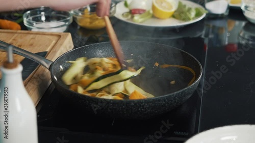 On stove there is frying pan with chopped carrots, zucchini, steam is coming out, vegetables stewed, cook adds olive oil, pours finely chopped onions and red pepper, mixes everything, wooden spatula photo