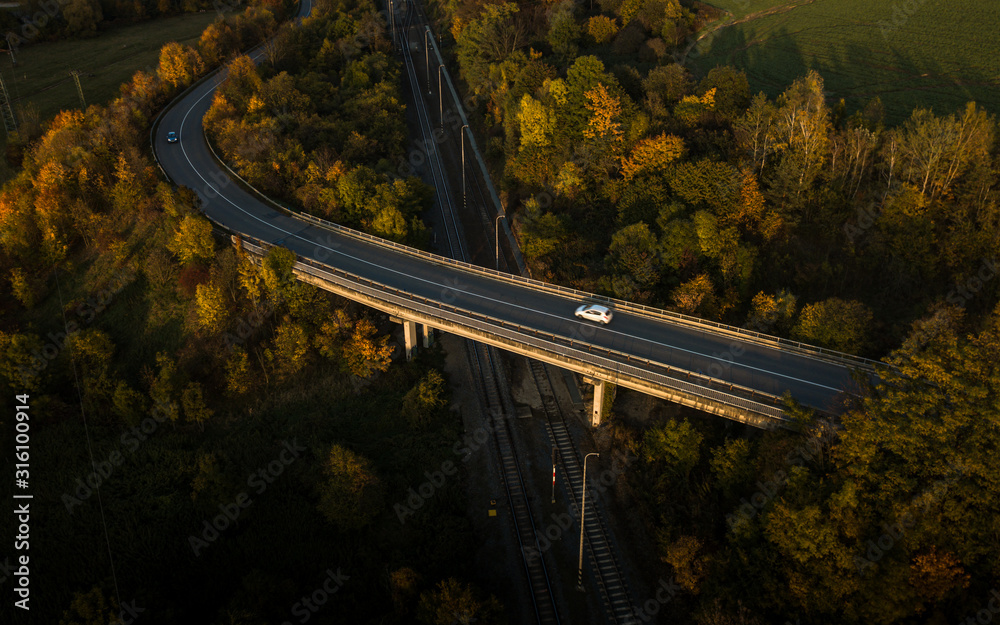 Winding road in autumn forest at sunset in mountains. Aerial view. Top view of beautiful asphalt roadway and orange trees. Highway through the woodland in fall. Trip in europe. Travel and nature - obrazy, fototapety, plakaty 