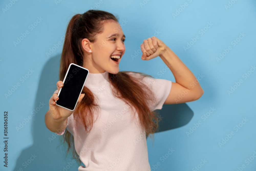 Showing phone screen, looks like winner. Caucasian teen girl's portrait on blue background. Beautiful model in casual wear. Concept of human emotions, facial expression, sales, ad. Copyspace. Happy. Showing phone screen, looks like winner. Caucasian teen girl's portrait on blue background. Beautiful model in casual wear. Concept of human emotions, facial expression, sales, ad. Copyspace. Happy.