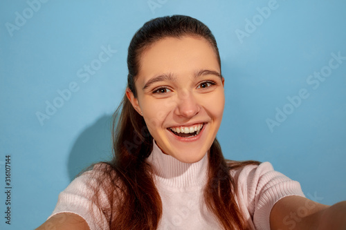 Posing for selfie taking. Caucasian teen girl s portrait on blue background. Beautiful model in casual wear. Concept of human emotions  facial expression  sales  ad. Copyspace. Looks happy.