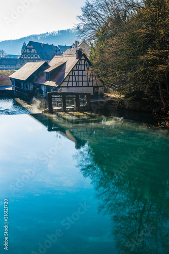 Germany, Early morning fog moving over water surface of blue pot or german blautopf in blaubeuren forest, a blue shiny natural source in warm sunlight on cold winter day