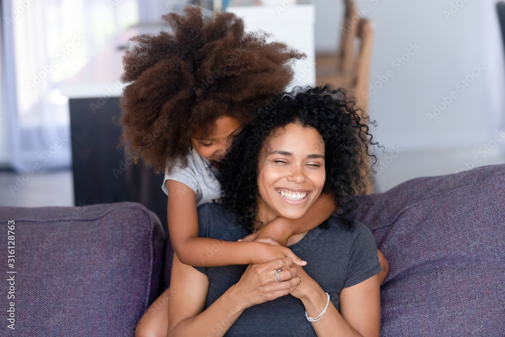 Little black girl hug smiling mom on couch from behind Little black girl hug smiling mom on couch from behind