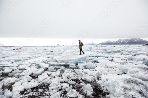 young male traveler in green clothes in a red hat and a green backpack stands on an iceberg on an ice lake in Iceland