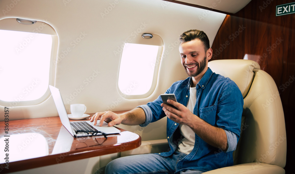 Perfect smile. Close-up photo of a good-looking man in a casual outfit, who is smiling, while typing something on his laptop and holding a smartphone in his left hand, flying first class of plane.