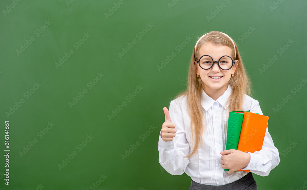 Smiling girl wearing funny glasses stands with books near a school chalkboard and shows thumbs up gesture. Empty space for text