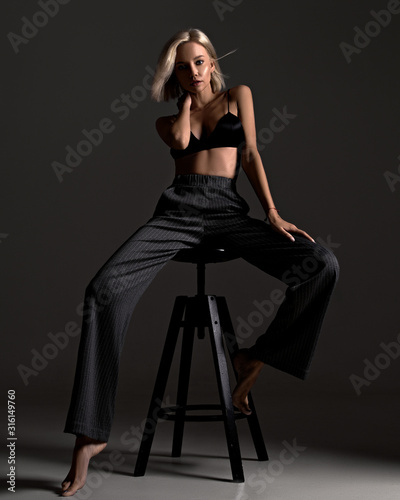 Stylish fashion elegant blonde woman model posing on black background in underwear and classic trousers. Sexy girl with beautiful face and body portrait in lingerie in room