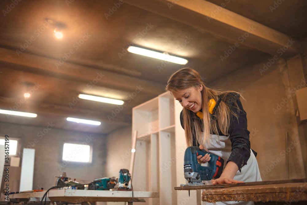 Attractive female carpenter using some power tools for her work in a woodshop. Young woman doing woodwork in a workshop. Woman craftswoman working in her workshop