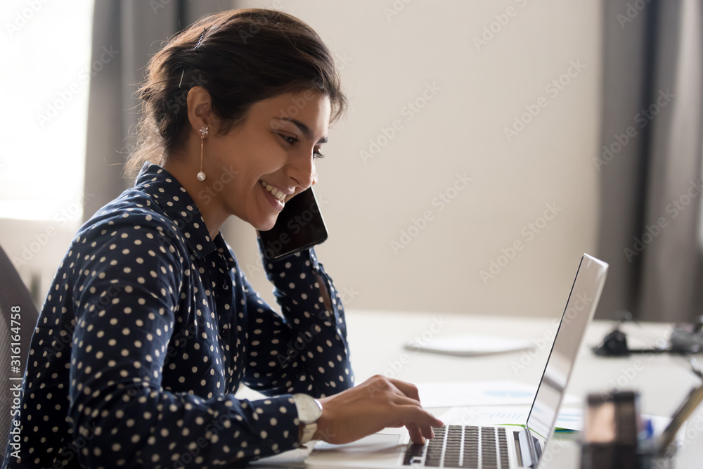 Smiling employee working on laptop talking over phone - obrazy, fototapety, plakaty 