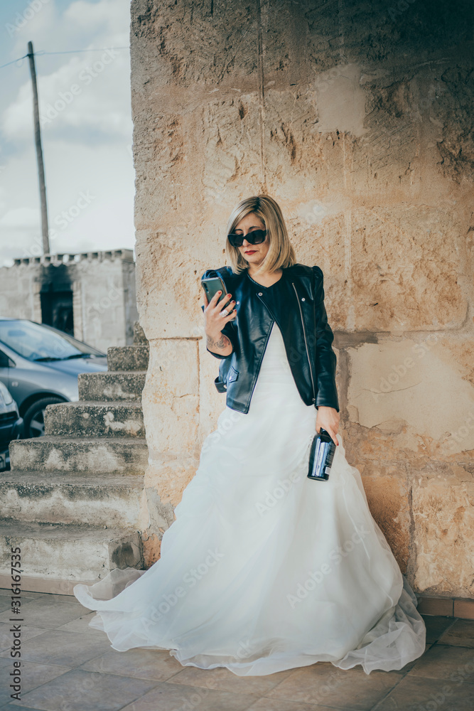 Foto Stock Disappointed attitude, blonde mature beautiful woman wearing a wedding  dress, sunglasses and a leather jacket, looking at a notification on her  smartphone while holding an alcohol bottle. Rocker look.