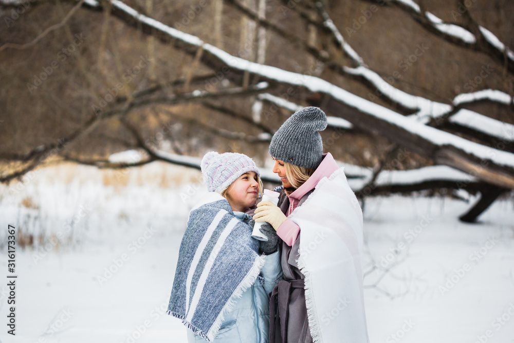 Mother and daughter are walking in the winter forest, wrapped in a blanket, tea or a drink from a thermos Mother and daughter are walking in the winter forest, wrapped in a blanket, tea or a drink from a thermos