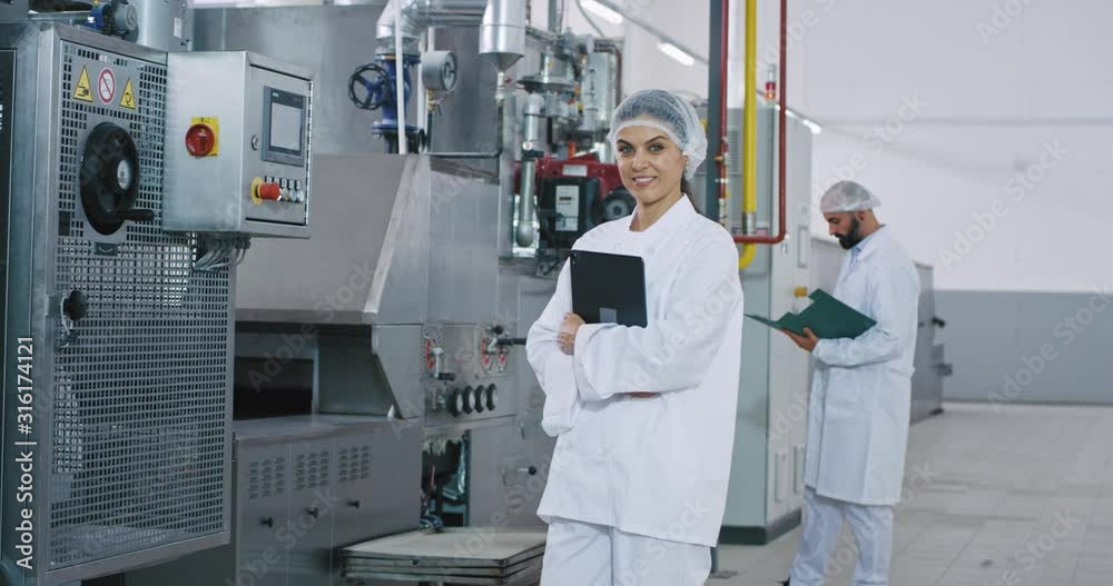 Good looking engineer woman with a perfect smile in a big manufacturing industry holding a electronic tablet looking straight to the camera and smiling background other engineer checking the Good looking engineer woman with a perfect smile in a big manufacturing industry holding a electronic tablet looking straight to the camera and smiling background other engineer checking the