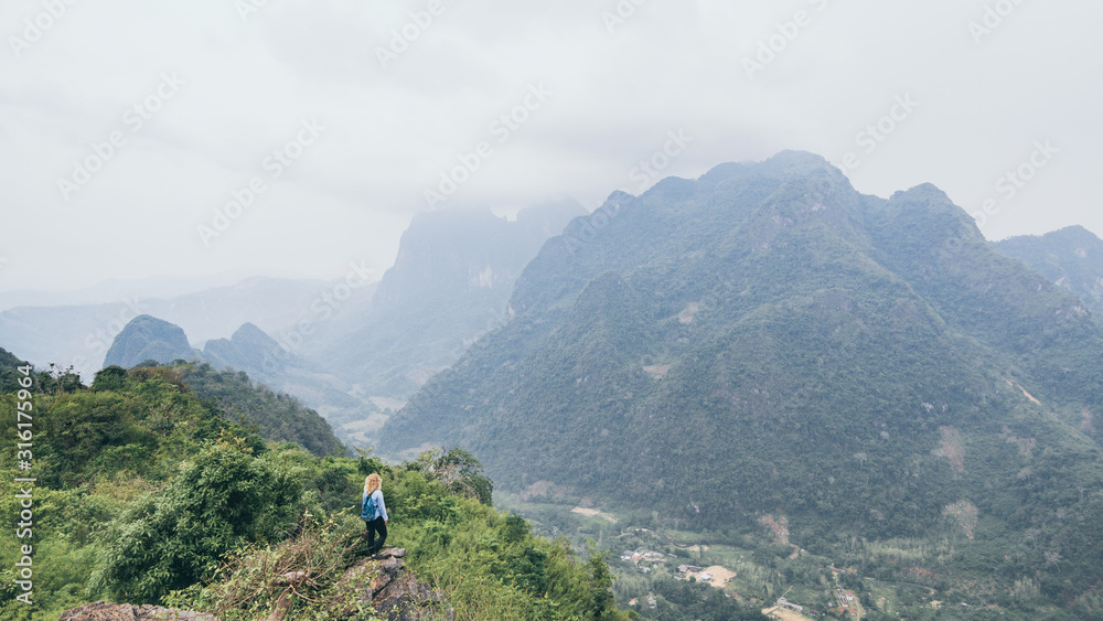 Young Caucasian woman standing on the top of the mountain overlooking river valley in Nong Khiaw village, Laos. Solo traveller with backpack