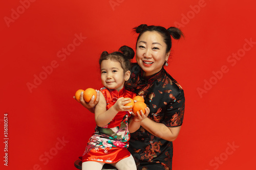 Hugging, smiling, holding mandarines. Happy Chinese New Year 2020. Asian mother and daughter portrait on red background in traditional clothing. Celebration, human emotions, holidays. Copyspace.