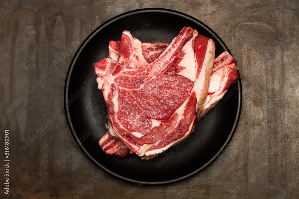 Chuletón de carne cruda de vaca sobre plato negro redondo. Vista superior