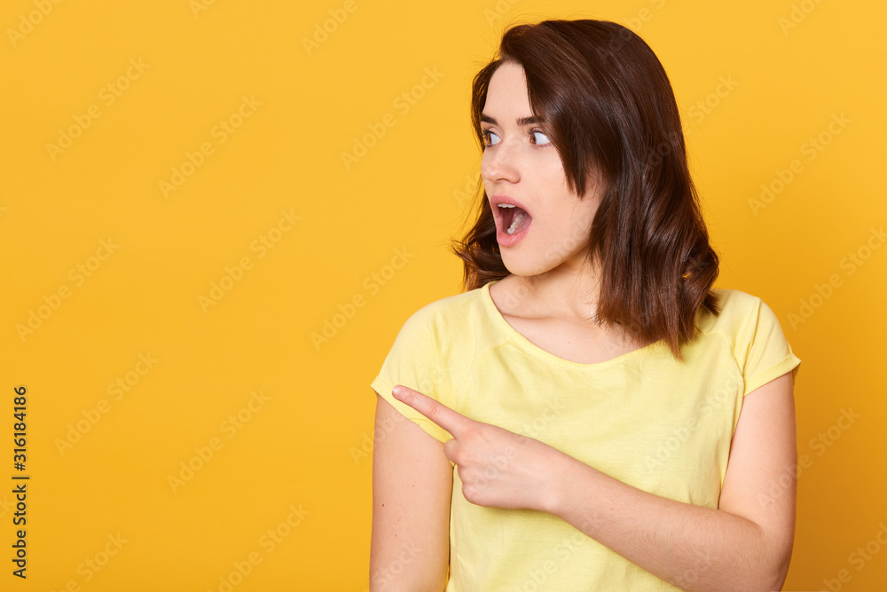 Close up portrait of young beautiful woman wearing casual t shirt, standing over isolated yellow background and points aside, looks worried and shocked. Copy space for advertisment or promotional text Close up portrait of young beautiful woman wearing casual t shirt, standing over isolated yellow background and points aside, looks worried and shocked. Copy space for advertisment or promotional text