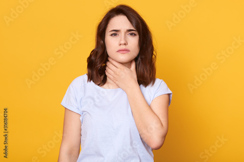 Close up portrait of cute sick young brunette woman wearing white casual t shirt, having sore throat, holding hand on her neck, sufferingfrom throat pain, painful swallowing, health care concept.