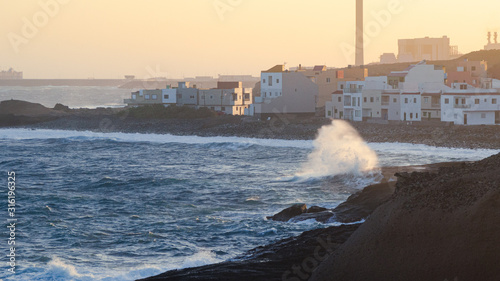 Waves hitting the rocky shore with houses and an industrial harbour with large chimney in the background, with copy space