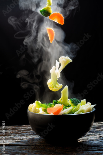 The steam from the vegetables carrot broccoli cauliflower on black bowl , a steaming. Boiled hot Healthy food on table on black background,hot food and healthy meal concept