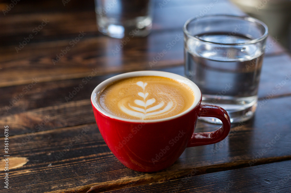 Water and red cup of coffee with latte art on wooden background. Table in cafe. Concept of easy breakfast. Small ceramic cup and glass Water and red cup of coffee with latte art on wooden background. Table in cafe. Concept of easy breakfast. Small ceramic cup and glass
