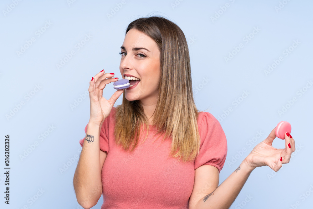Young blonde woman over isolated blue background holding colorful French macarons and eating it Young blonde woman over isolated blue background holding colorful French macarons and eating it