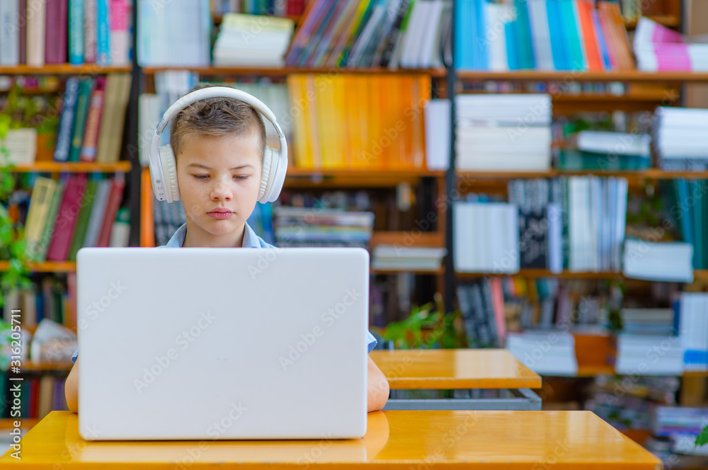 Cute boy in the library with headphones working on a laptop, focusing, looking at the screen Cute boy in the library with headphones working on a laptop, focusing, looking at the screen