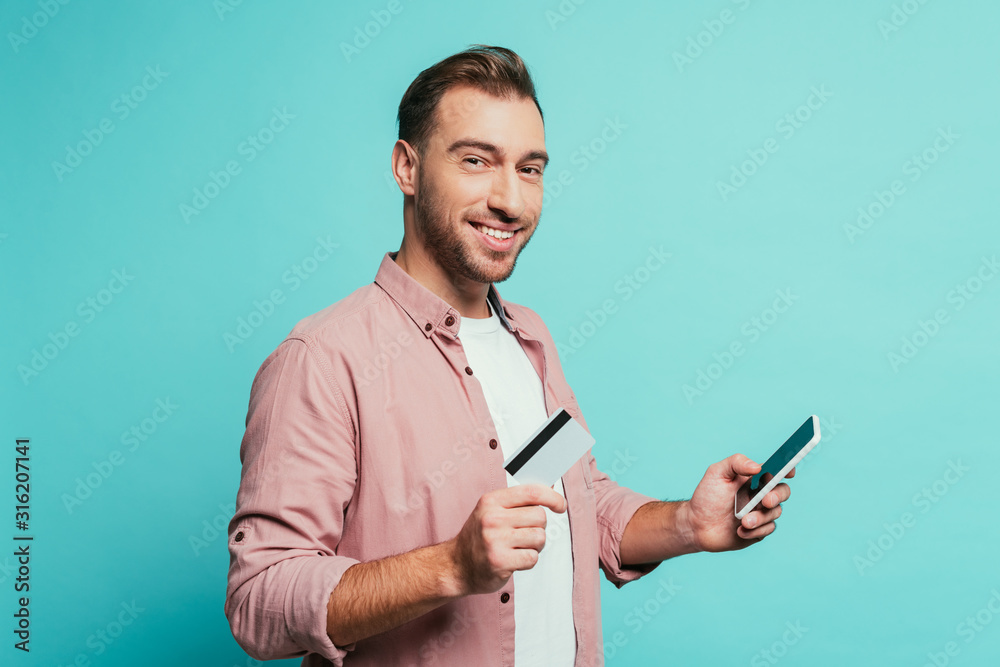 cheerful bearded man shopping online with smartphone and credit card, isolated on blue