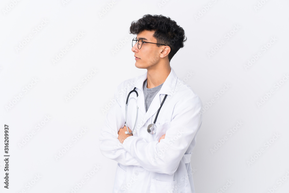 Young Argentinian man over isolated white background wearing a doctor gown and with arms crossed looking side Young Argentinian man over isolated white background wearing a doctor gown and with arms crossed looking side